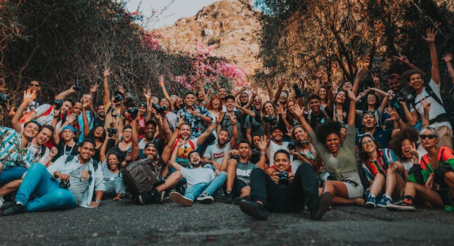 Group of multiracial happy people sitting with raised hands on asphalt road near mountains and trees in summer for portrait photo and looking at camera
